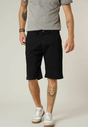 Homme portant un short noir jusqu'aux genoux, un t-shirt gris, des baskets blanches et des bracelets, avec des tatouages sur les bras et les jambes sur un fond uni.