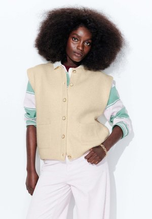 Gilet en laine beige avec un devant à boutons et deux poches, superposé à une chemise à rayures multicolores. Le mannequin porte un pantalon en velours côtelé rose clair.