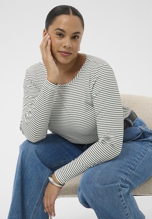 Kvinde med mørkt hår i en lav knold iført en hvid og sort stribet langærmet trøje og blå jeans, der sidder og hviler sit ansigt på sin hånd.
