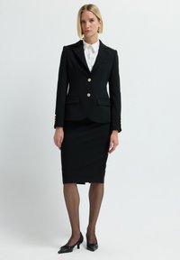 Completo giacca nero su misura con giacca aderente adornata da bottoni dorati, abbinata a una gonna a tubino fino al ginocchio. Camicia bianca sotto, décolleté neri.