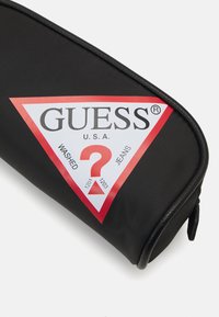 Svart kosmetikapåse med en triangulär vit och röd logotyp som visar "GUESS U.S.A." och "WASHED JEANS." Har dragkedjestängning och rundade kanter.