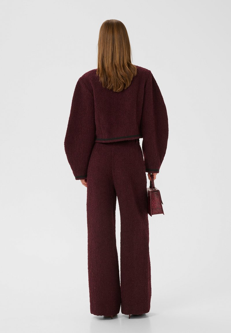 Burgunderfarbenes Zweiteiler-Outfit mit einer verkürzten, strukturierten Jacke mit Puffärmeln und weitem Hosenbein, ergänzt durch eine kleine Handtasche.