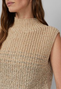 Frau in einem ärmellosen beigen gestrickten Pullover mit hohem, geripptem Kragen, von den Schultern bis zum Mund gezeigt, mit langen braunen Haaren.