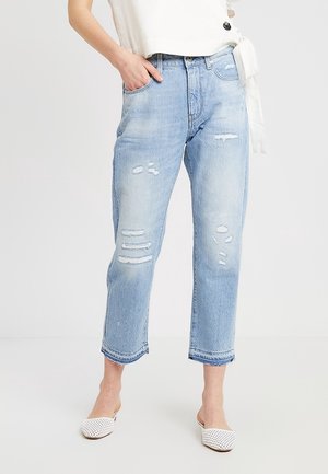 Žena má na sobě světle modré roztrhané zkrácené džíny s třepícími se lemy a bílé pletené slip-on boty, stojí s jednou rukou v kapse.