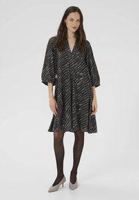 Schwarzes Kleid mit V-Ausschnitt und Ballonärmeln, das ein welliges weißes Muster aufweist. Dazu werden transparente schwarze Strumpfhosen und hellgraue spitze Absätze getragen.