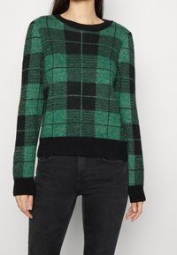 Pull à carreaux verts et noirs avec un col rond, des poignets et un ourlet côtelés. En tissu texturé. Accents noirs sur les bords.