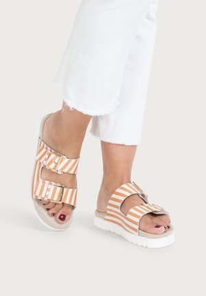Feet wearing orange and white striped slide sandals with double gold buckles and white soles, paired with cropped white pants.