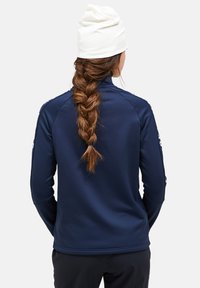 Marineblaue Zip-Jacke mit langen Ärmeln und hohem Kragen, mit glatter Textur und Logodetail am Ärmel. Weiße Mütze getragen.