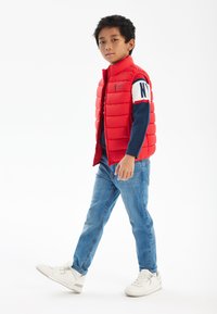 Röd vadderad väst med hög krage, bärs över en marinblå långärmad tröja. Ljust blåjeans och vita sneakers fulländar outfiten.