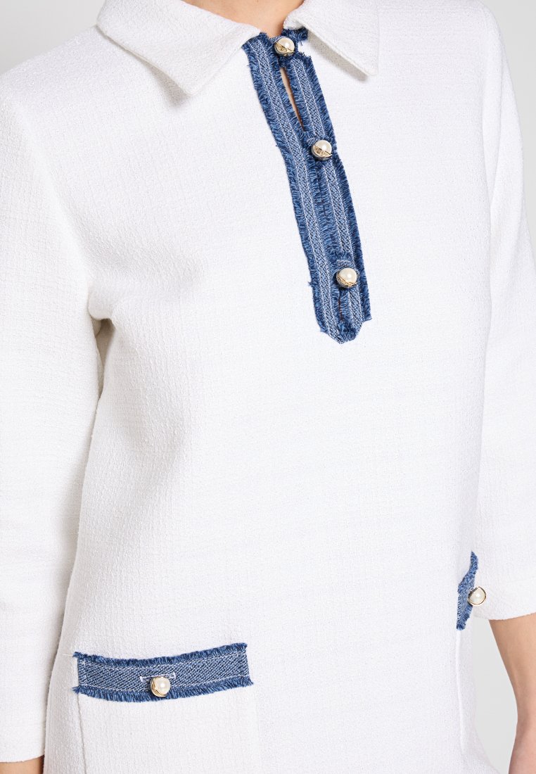 Weißes Tunika-Shirt mit strukturiertem Finish, versehen mit blauen Denim-Akzenten am Kragen und an den Taschen, geschmückt mit runden metalen Knöpfen.