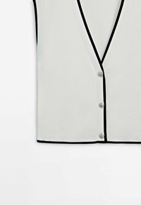 Cardigan blanc sans manches avec bordure noire, bords côtelés, col en V et trois boutons argentés sur le devant.
