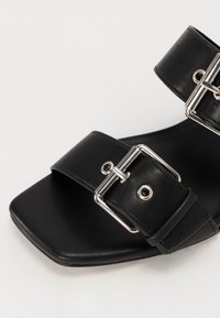 Sandale en cuir noir avec bout carré et deux brides à boucles argentées sur le pied et la cheville, sur fond uni.