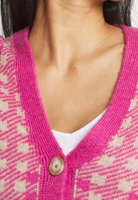 Růžový pletený cardigan s výstřihem do V a hvězdicovým vzorem, který má velký béžový knoflík na výstřihu a texturovaný, jemný materiál.