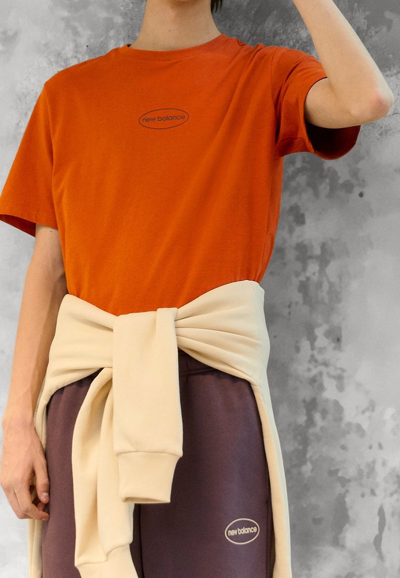 Oranje T-shirt met korte mouwen en een zwart logo. Beige trui om de taille gebonden, gecombineerd met een bruine broek met logo.
