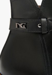 Schwarze Leder-Stiefelette mit V-Ausschnitt, genähten Details und einem silbernen Hardware-Akzent mit dem "NG"-Logo am Riemen.