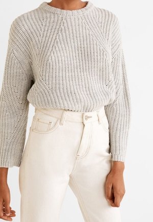 Pull en maille épaisse gris clair à manches longues associé à un pantalon taille haute écru sur une personne debout devant un fond blanc.