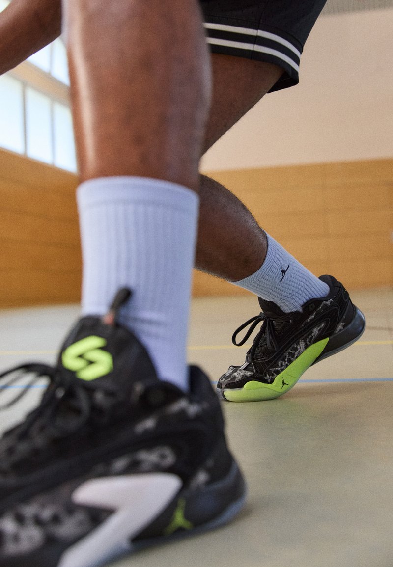 Zapatillas negras con acentos verdes, con detalles de superficie texturizada y cordones. Llevadas con calcetines deportivos de color púrpura claro y con relieve en un gimnasio.