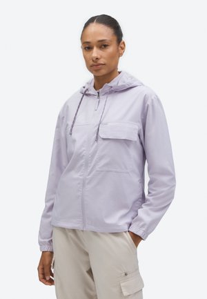 Naine kannab helelillat lukuga kapuutsiga jopet, millel on rinnatasku, ja beežikaid pükse, seistes ühe käega taskus.