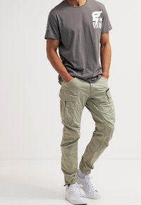 Šedé tričko s bílým potiskem na přední straně, nošené s světle zelenými kalhotami typu cargo s několika kapsami a elastickými manžetami, doplněné bílými teniskami.