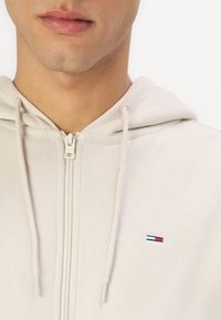 Svijetlosmeđa zip-up majica s kapuljačom i vezicama, sa malim izvezenim logotipom u plavoj, crvenoj i bijeloj boji na lijevoj strani prsnog dijela.