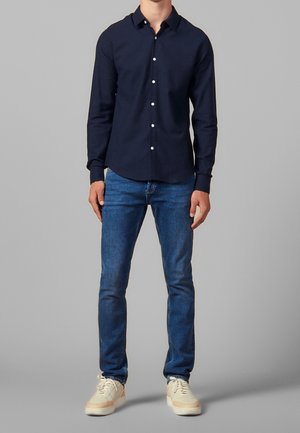 Mann trägt ein tailliertes, navyfarbenes Hemd mit Knöpfen, blaue schmale Jeans und weiße Sneaker, steht vor einem grauen Hintergrund.