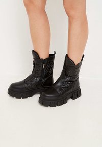 Botines negros hasta el tobillo con un patrón acolchado, fabricados con material similar al cuero, que cuentan con una suela gruesa y cremalleras laterales para facilitar su uso.