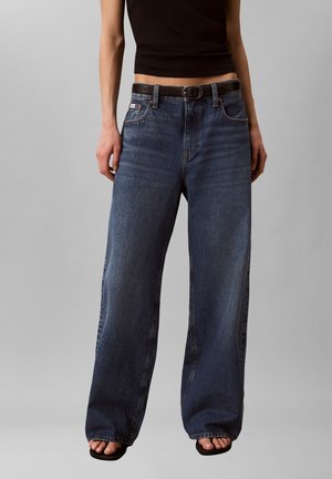 Slank bovenlichaam met een zwart cropped tanktop, wijd uitlopende donkerblauwe spijkerbroek met zwarte riem en zwarte open-toe sandalen op een grijze achtergrond.