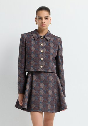 Woman wearing a brown-blue patterned jacket with large gold buttons and a matching flared skirt, standing against a plain white background.