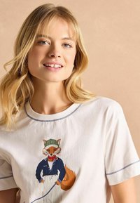 Valge puuvillane T-särk, millel on rebane illustratsioon, seljas sinine särk ja peas roheline müts, ning kontrastne sinine õmblus kaelaaugu ümber.