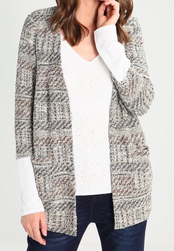 Grau-weiße Strickjacke mit strukturiertem Muster, offenem Vorderteil und lockeren Ärmeln, über einem weißen Oberteil mit dezenten Mustern getragen.