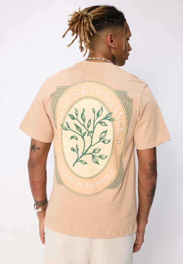 T-shirt à manches courtes couleur pêche avec un grand motif botanique vert dans le dos, accompagné du texte "FLEURS & FRAGRANCE JARDIN DE PROVENCE".