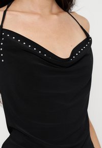 Femme portant un haut noir sans manches avec de fines bretelles et des clous argentés le long de l'encolure.
