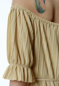Hellgelbes Off-Shoulder-Oberteil mit vertikalen Streifen, Pufärmeln, gerafftem Ausschnitt und ausgestellten Manschetten. Glatte Textur, leichtes Material.