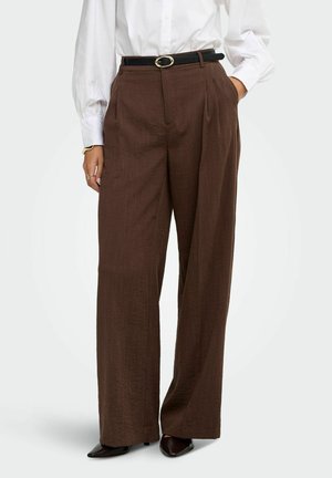 Femme portant un pantalon marron taille haute à jambes larges avec une ceinture noire, une blouse blanche et des chaussures pointues foncées, debout les mains dans les poches.