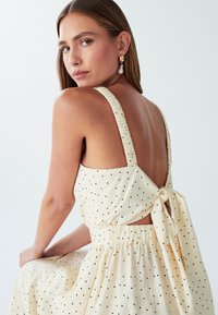 Vestido beige claro con lunares negros, tirantes texturizados, espalda abierta y cintura atada. Suave tela y silueta fluida mejoran la comodidad.