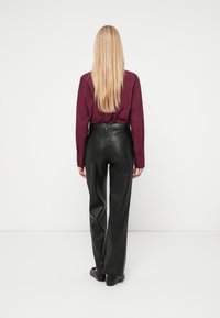 Musta nahkainen korkeavyötäröinen housu suoran lahkeen muotoilulla. Pitkähihainen burgundy-paita. Malli nähtynä takaa, suorilla vaaleilla hiuksilla.