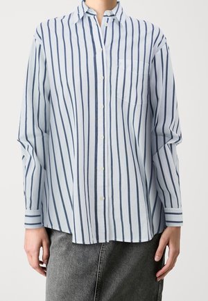 Osoba ubrana w białą koszulę na guziki z pionowymi granatowymi paskami i kieszenią na piersi, w zestawieniu z ciemnoszarymi dżinsami.