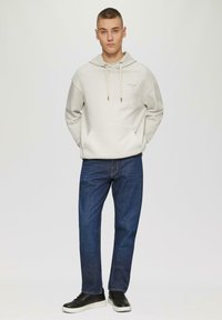 Hoodie gris clair avec poche avant, cordons de serrage et impression subtile. Associé à un jean bleu foncé et des baskets noires avec des semelles blanches.