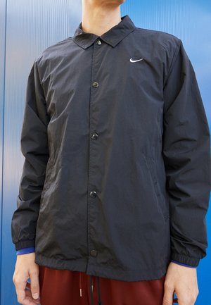 Person iført en sort, knappet Nike-jakke med elastiske manchetter og røde bukser foran en blå baggrund.