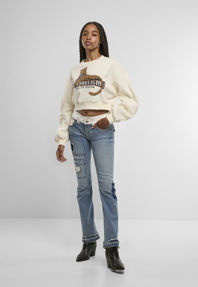 Sweatshirt court crème avec des détails en imprimé léopard, associé à un jean bleu clair avec des patchs et des ourlets effilochés. Bottes à talons noires.