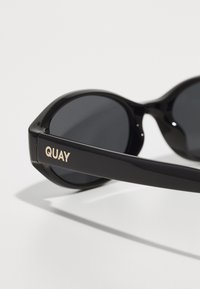 Lunettes de soleil noires avec des verres ronds, une monture brillante et une branche rectangulaire portant le nom de la marque "QUAY" en doré.