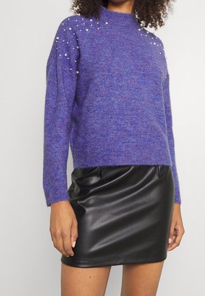 Femme portant un pull en tricot violet avec des perles décoratives sur les épaules et une mini-jupe en cuir noir, debout devant un fond uni.