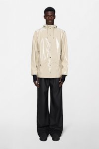 Μπεζ, γυαλιστερό αδιάβροχο με κουκούλα, μπροστινό κούμπωμα με κουμπιά και πλαϊνές τσέπες, συνδυασμένο με χαλαρά μαύρα παντελόνια και μαύρα παπούτσια.