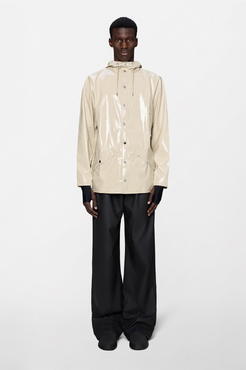Μπεζ, γυαλιστερό αδιάβροχο με κουκούλα, μπροστινό κούμπωμα με κουμπιά και πλαϊνές τσέπες, συνδυασμένο με χαλαρά μαύρα παντελόνια και μαύρα παπούτσια.
