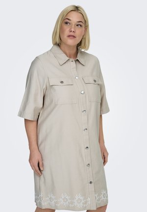 Blonde Frau in beigem Hemdblusenkleid mit kurzen Ärmeln, Brusttaschen und weißer Blumenstickerei am Saum, die nach links schaut.