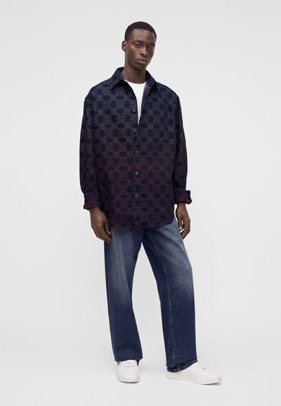 Homme portant une chemise boutonnée sombre à motifs, jean bleu, baskets blanches, debout devant un fond clair uni.