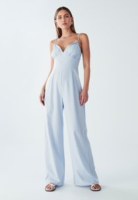 Mujer vestida con un mono de pierna ancha de color azul claro con tirantes finos y sandalias blancas de punta abierta, de pie contra un fondo blanco liso.