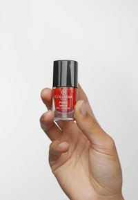 Lacca per unghie rossa in una bottiglia di vetro con un tappo nero; l'etichetta riporta "COLLISTAR PURO SMALTO NAIL LACQUER." Tenuta da una mano su uno sfondo bianco.