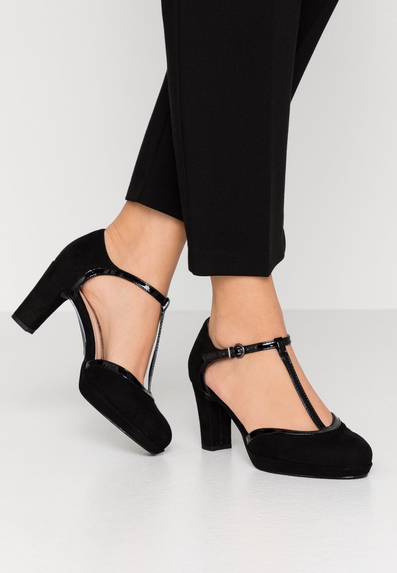 Pieds portant des chaussures à talons hauts en suède noir à bride en T, associées à un pantalon noir court, sur un fond clair uni.