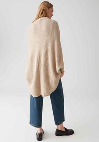 Femme portant un poncho texturé beige, un jean court bleu et des mocassins noirs, se tenant devant un fond gris clair uni.